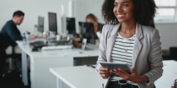 Happy thoughtful young businesswoman with digital tablet in hand smiling and looking away in front of colleague at background