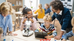 Elementary School Science Classroom: Cute Little Girl Looks Under Microscope, Boy Uses Digital Tablet Computer to Check Information on the Internet. Teacher Observes from Behind, STEM education, gender neutral