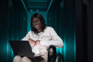 Portrait of disabled African-American woman using laptop and looking at camera while working in server room, accessible job opportunity, copy space